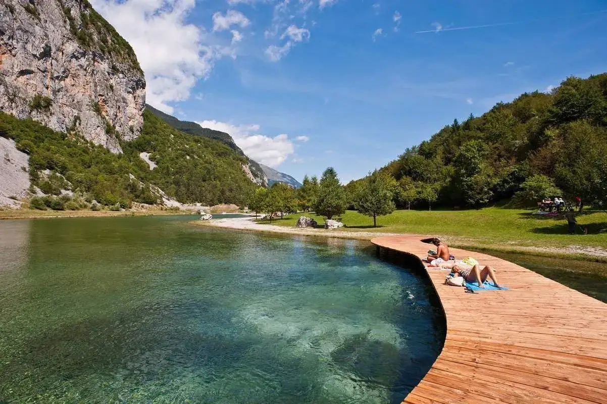 Passeggiata sul pontile in legno dei Bagni di Rabbi, con persone che prendono il sole e un lago cristallino circondato da montagne verdi.