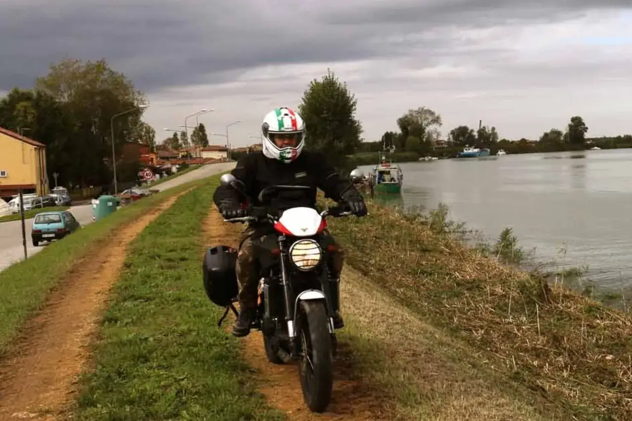 Motociclista esplora il delta del Po, con barche sul fiume e un cielo nuvoloso.