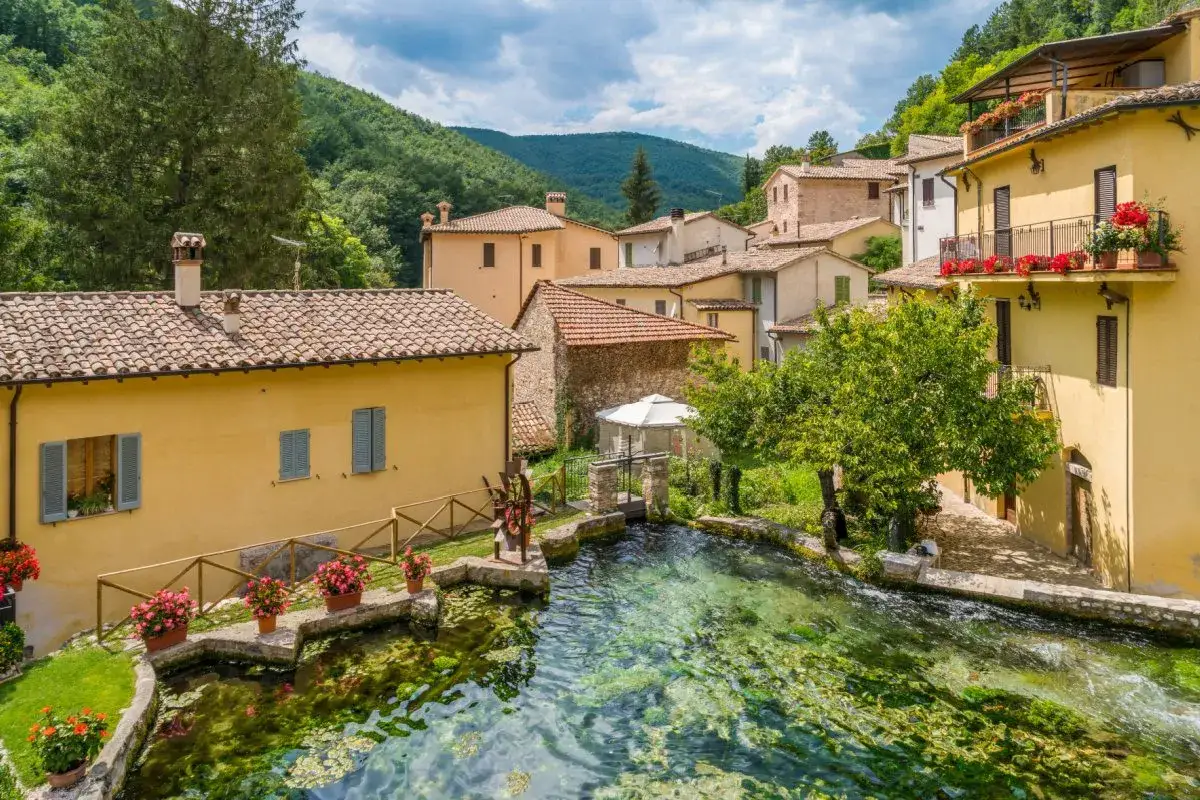 Un borgo incantevole, la Venezia dell'Umbria, con case gialle affacciate su un limpido ruscello, un mulino e fiori rossi.