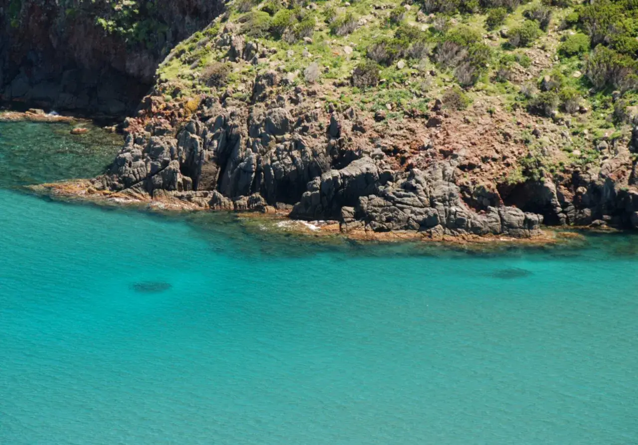 Scogliera selvaggia e mare turchese a Capraia. L'acqua cristallina lambisce rocce rosse e verdi macchie mediterranee.