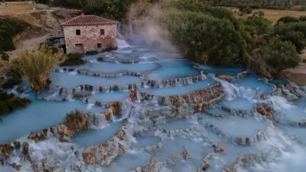 Cascate naturali con acqua azzurra e vapore, un casolare in pietra e due persone che si godono le terme di Saturnia.