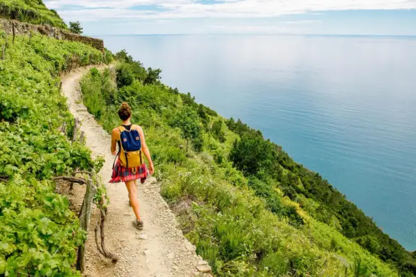 Donna con zaino cammina sul sentiero dell'Alta Via dei Monti Liguri, con il mare azzurro a destra e vigneti a sinistra.