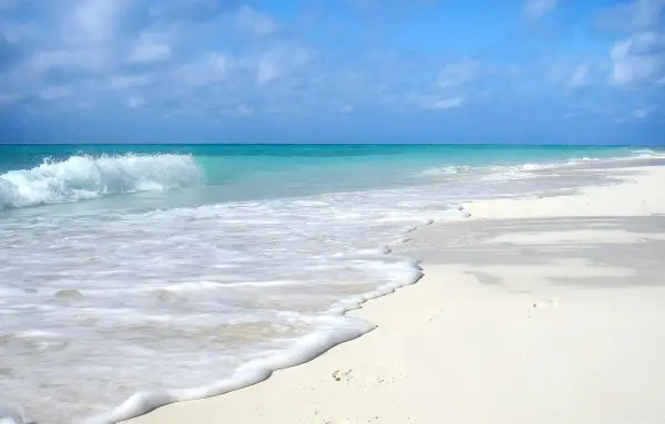 Onda che lambisce la sabbia bianca di una spiaggia d'inverno, con cielo azzurro e nuvole sparse.
