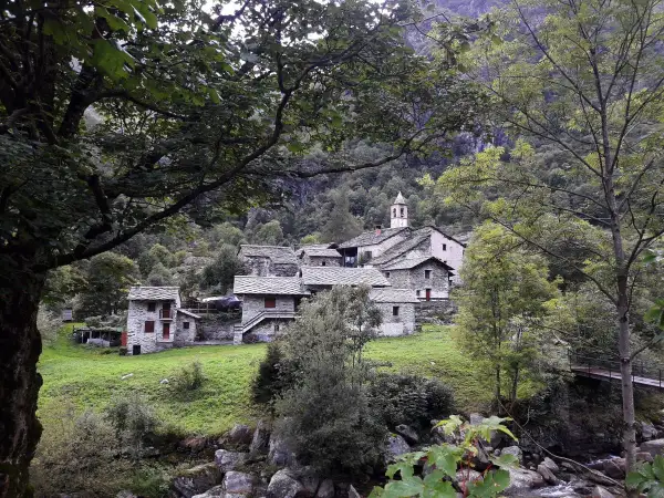 Borgo in pietra con campanile, immerso nel verde della Val Chiuse lla. Un ponte attraversa il torrente.