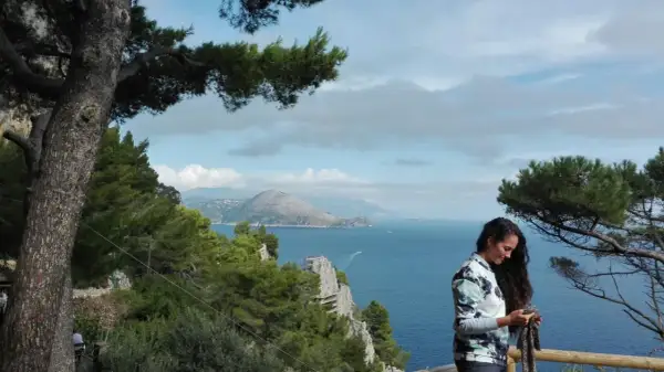 Donna con sciarpa guarda il cellulare, con vista mare e montagne di Capri a piedi.