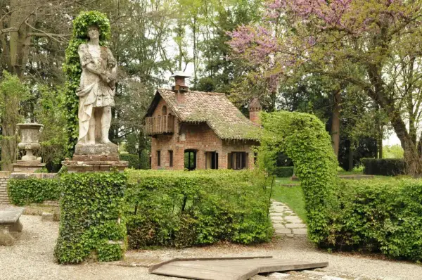 Un giardino incantato a Grazzano Visconti, dove si trova un'antica statua e una casetta in mattoni circondata da siepi verdi.