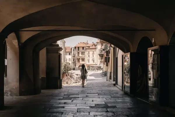 Una donna cammina sotto un portico verso una piazza soleggiata, con biciclette parcheggiate e palazzi storici che ricordano i paesi delle Langhe.