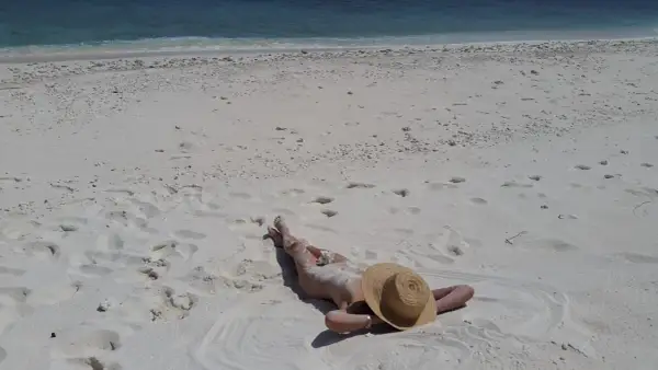 Person lying on a white sandy beach, wearing a straw hat, with the blue ocean in the background.