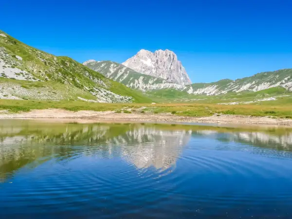 Specchio d'acqua riflette le maestose montagne del centro Italia, con verdi pendii e un picco roccioso sotto un cielo azzurro.