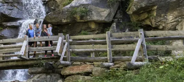 Famiglia felice sul sentiero delle cascate, con una cascata fragorosa alle spalle e un ponte di legno rustico.