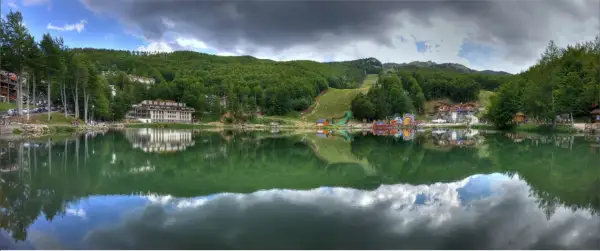 Lago alpino con riflessi di alberi e case, sullo sfondo il **passo del Cerreto** con piste da sci e giochi per bambini.