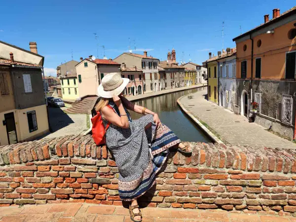 Donna con cappello di paglia e zaino rosso seduta su un ponte di mattoni a Comacchio, con canali e case colorate. Un luogo ideale per una gita con bambini.