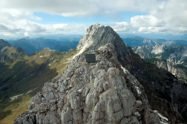 Bivacco Luca Vuerich, un cubo scuro su una cresta rocciosa, domina un panorama alpino mozzafiato sotto un cielo nuvoloso.