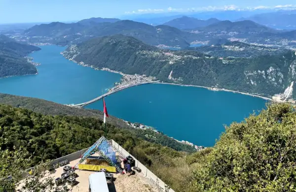Vista panoramica del Lago di Lugano, con un ponte che lo attraversa. In primo piano, una struttura per eventi e una bandiera. Ideale per le val d'Intelvi passeggiate.