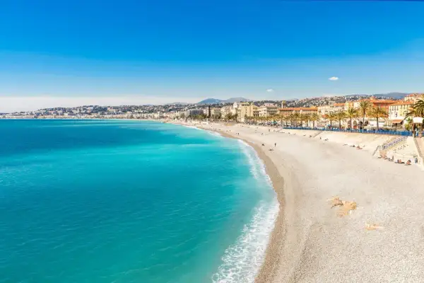 Spiaggia di ciottoli baciata dal sole sulla costa azzurra, con il mare turchese che lambisce la riva e palme che ombreggiano la Promenade des Anglais.