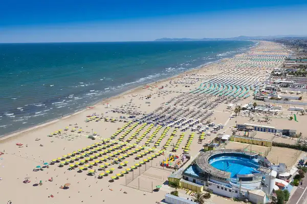 Vasta spiaggia della **costiera romagnola** con file ordinate di ombrelloni gialli e azzurri, mare calmo e un complesso con piscina.