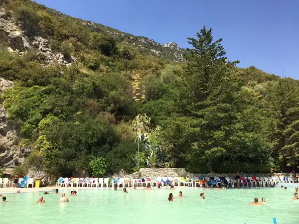 Piscina con persone che nuotano, circondata da vegetazione lussureggiante e rocce, un luogo che sembra uscito dalle grotte delle ninfe.
