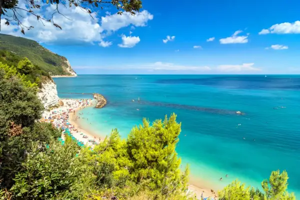 Spiaggia affollata con ombrelloni colorati e gente che nuota nelle acque cristalline. Una scogliera bianca domina la costa, incorniciata dal verde della vegetazione. Una perfetta località mare Marche.