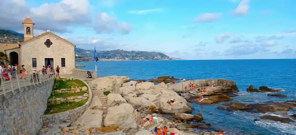 La chiesa bianca domina la costa rocciosa di Bordighera, con bagnanti che si godono il sole. Un luogo incantevole per scoprire cosa vedere a Bordighera.