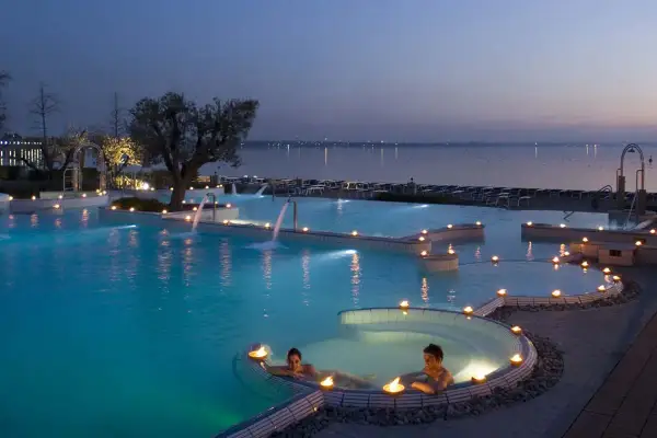 Coppia rilassata in una piscina termale illuminata da candele, con cascate e vista lago al tramonto. Un'oasi di pace alle terme a Brescia.