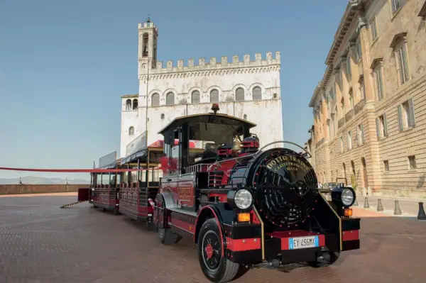 Il trenino turistico di Gubbio, rosso e nero, attende i passeggeri in una piazza storica con palazzi antichi.
