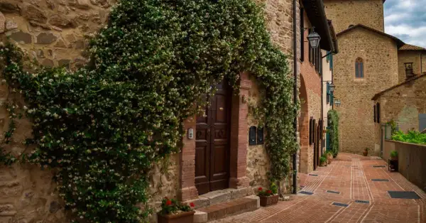 Un antico borgo in Umbria, con un portone di legno scuro avvolto da rampicanti fioriti. La strada in mattoni rossi sale dolcemente tra case in pietra.