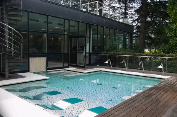 Piscina moderna con mosaico azzurro e bianco, scala a chiocciola in metallo, vetrate e alberi rigogliosi. Un'oasi di relax a Boario Terme.