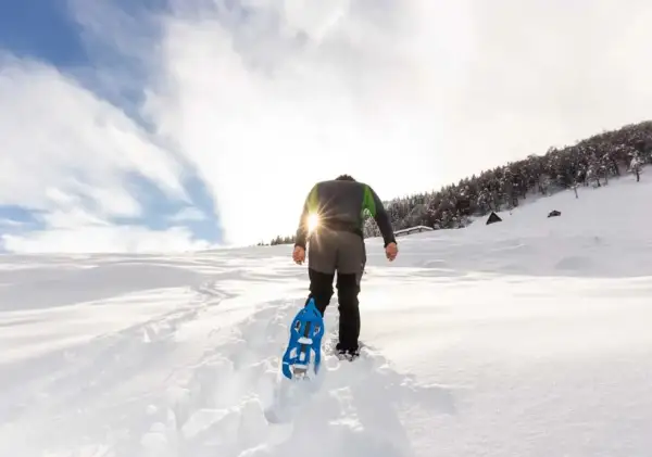 Escursionista con racchette da neve cammina su sentiero innevato, scoprendo posti belli in montagna Piemonte inverno. Sole splendente tra le nuvole.