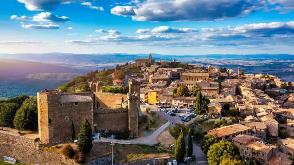 Scorci borghi antichi arroccati su una collina, con un castello imponente e case in pietra sotto un cielo azzurro con nuvole bianche.