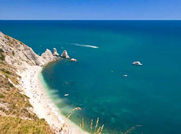 La spiaggia più bella del Conero, con sabbia bianca, mare cristallino e scogliere imponenti. Barche solcano le acque turchesi.