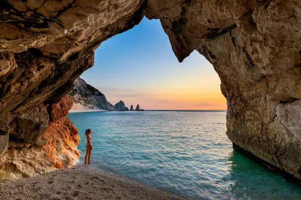 Vista da una grotta sulle spiagge della Riviera del Conero al tramonto. Una ragazza ammira il mare turchese e le rocce a picco.