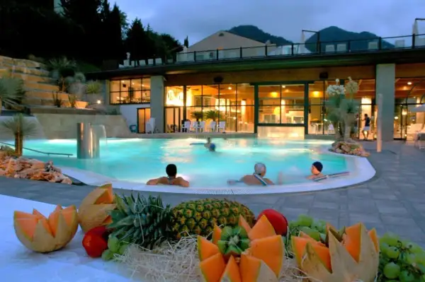 Piscina termale illuminata di notte, con persone che nuotano e un tavolo di frutta fresca in primo piano. Un'oasi di relax che ricorda le terme di Reggio Emilia.