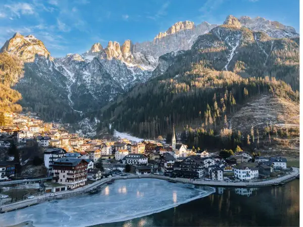 Villaggio alpino innevato ai piedi delle Dolomiti, ideale per escursioni invernali. Lago ghiacciato riflette le montagne.