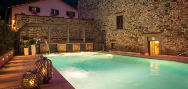 Piscina termale illuminata da lanterne, con cascate d'acqua e un edificio in pietra. Un'oasi di relax che ricorda le terme della Riviera Romagnola.