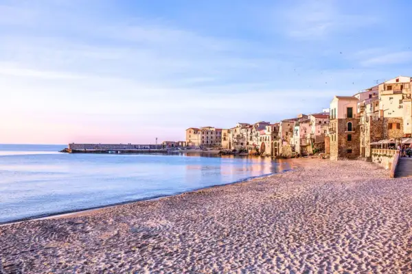 La spiaggia di Cefalù al tramonto, con le case colorate che si affacciano sul mare calmo e la sabbia dorata.